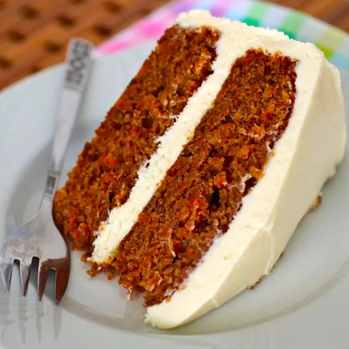 A slice of two-layer carrot cake topped with cream cheese frosting, served on a white plate with a fork on the side