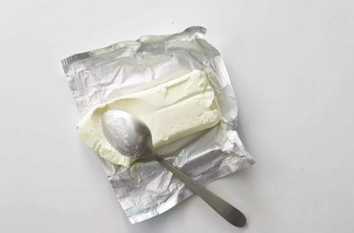 Block of cream cheese wrapped in aluminum foil with a spoon beside it