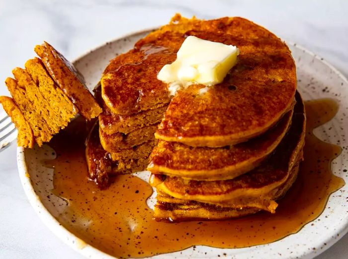 A high-angle shot of five stacked pumpkin pancakes on a speckled white plate, topped with a pat of butter and drenched in syrup.