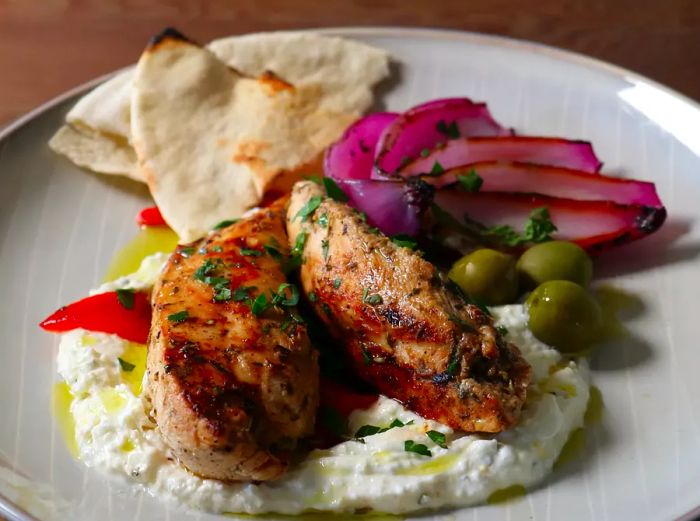 Chef John's Grilled Greek Chicken Breasts with Whipped Feta served on a plate with olives, grilled red onion, and pita in the background