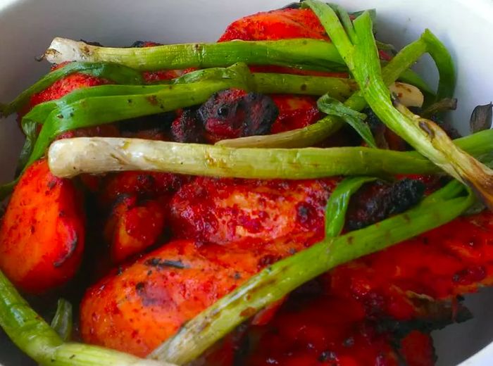 close-up of reddish-orange grilled chicken thighs (Chef John's Tandoori Chicken), topped with grilled green onions, served in a white bowl