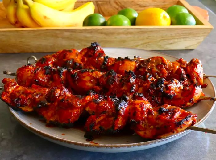 three perfectly grilled skewers of Chef John's Filipino-Style Barbecue Chicken, slightly charred, served on a platter with a wooden tray of bananas and citrus fruits in the background