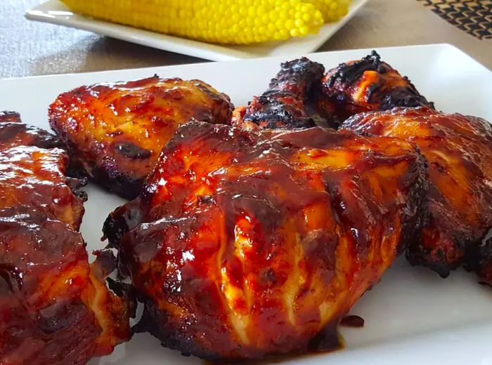 a white ceramic platter showcasing grilled chicken, beautifully coated with sticky barbecue sauce, accompanied by corn on the cob in the background