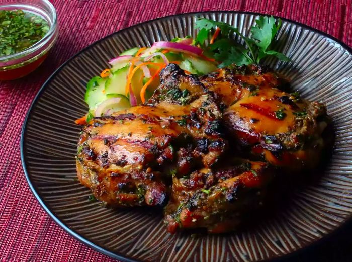 a plate of grilled chicken thighs topped with herbs, accompanied by a dipping sauce in the background