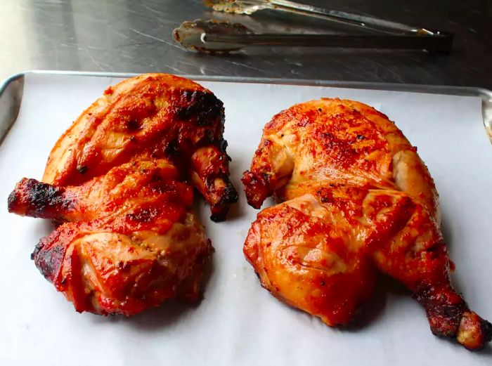 two grilled chicken halves (Chef John's Miso Honey Chicken) on a parchment-lined sheet pan, with a pair of tongs resting in the background