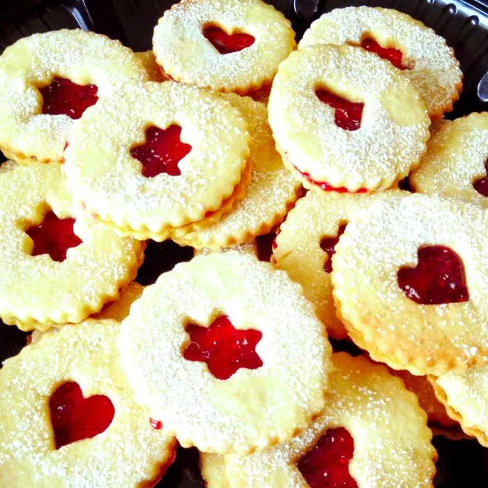 An overhead shot of homemade Cranberry Cornmeal Linzer sandwich cookies, filled with cranberry jelly, topped with cutout designs, and dusted with powdered sugar.