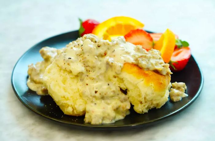 A plate of savory sausage gravy served over a fluffy biscuit, paired with fresh fruit on the side.