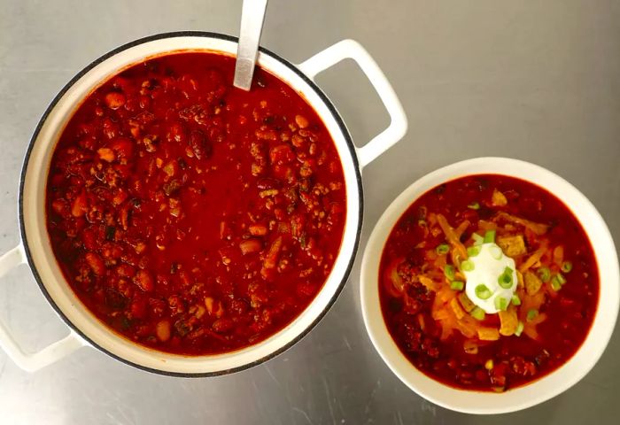 A top-down view of a simmering pot of chili beside a bowl garnished with shredded cheese, crunchy corn chips, fresh scallions, and a dollop of sour cream.