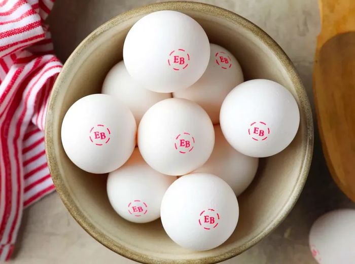 A bowl of Eggland's Best eggs resting on the counter, accompanied by a red striped towel and a wooden spoon.