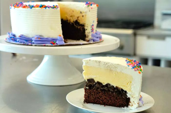 A side view of a homemade ice cream cake slice featuring a chocolate cake base, crunchy cookie layer, vanilla ice cream top, and a whipped cream frosting decorated with colorful candy sprinkles.