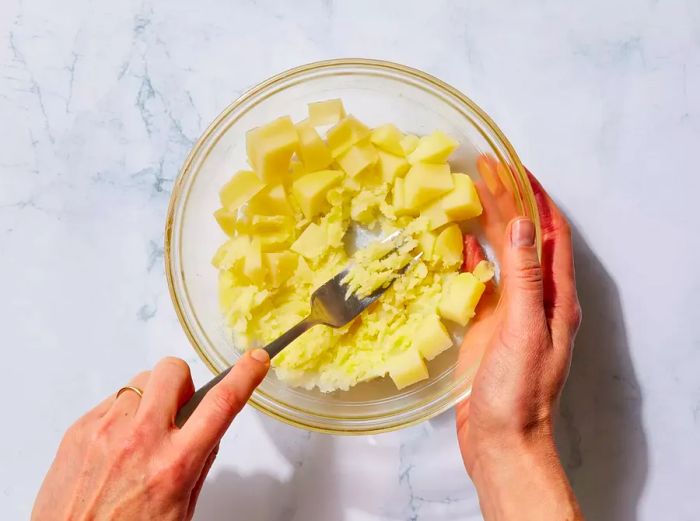 A glass bowl with mashed potatoes being mashed with a fork.