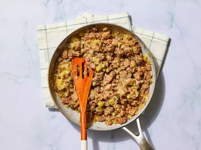 A pan of seasoned, browned ground meat being combined with mashed potatoes using a wooden spatula.