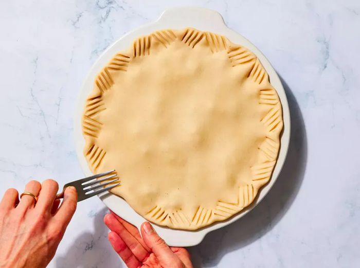 A top pie crust being pressed down along the edges with a fork.