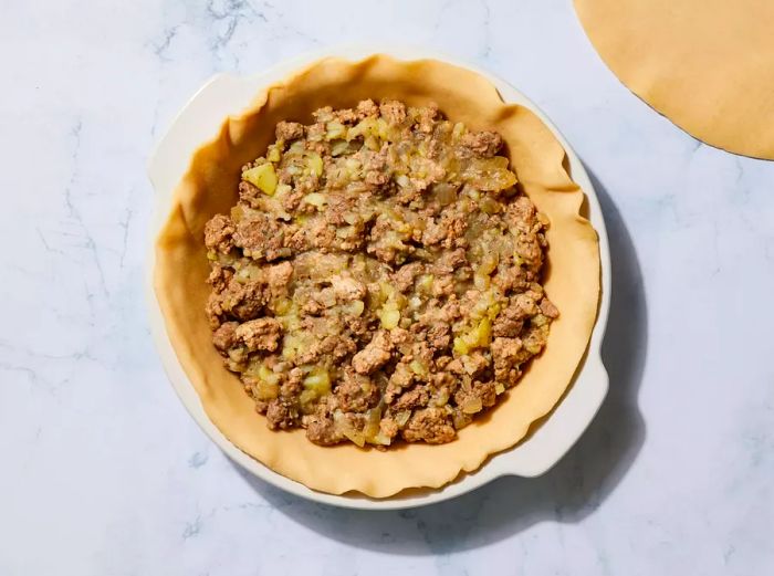 A pie dish lined with dough and filled with a ground meat and potato mixture.