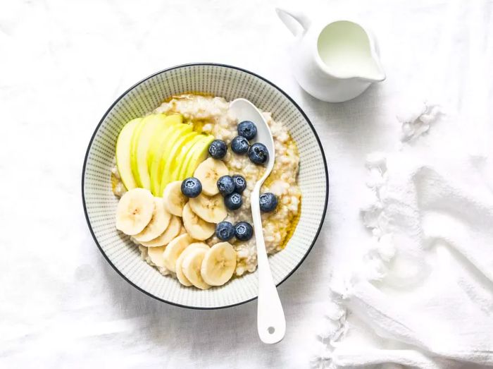 Oats breakfast porridge made with coconut milk, topped with apple, banana, blueberries, and a drizzle of honey on a light background, viewed from above. A healthy vegetarian option.