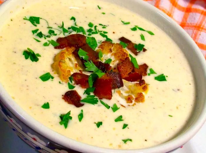 A close-up of Roasted Cauliflower Soup topped with cauliflower, crispy bacon, and fresh herbs, served in a bowl