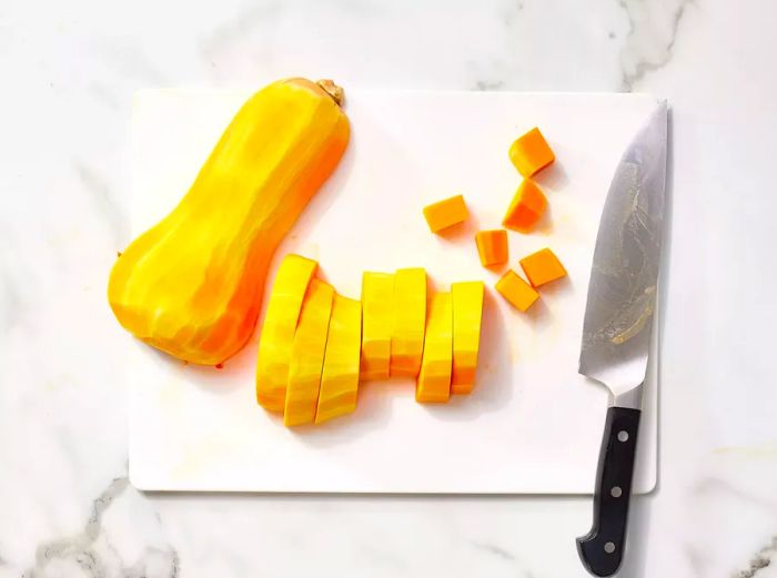 A cutting board with a peeled butternut squash, sliced into rounds and cubed