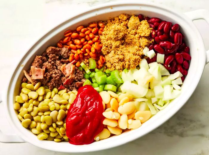 A large baking dish filled with beans, browned ground beef and bacon, brown sugar, celery, ketchup, and diced onions