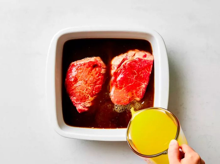 Pouring the orange marinade over raw steaks in a white baking dish.