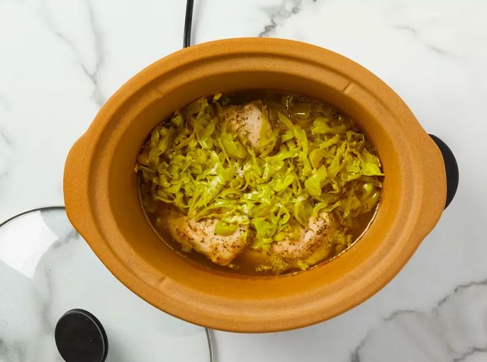 A top-down view showing pepperoncini peppers, butter, and 1/2 cup of reserved pepper juice placed over chicken in a crockpot.