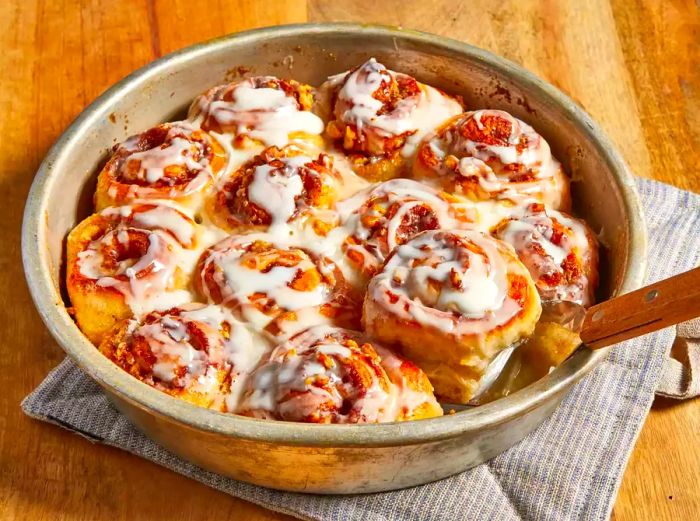 Close-up of freshly baked cinnamon rolls with icing in a round dish, with a spatula beside them.