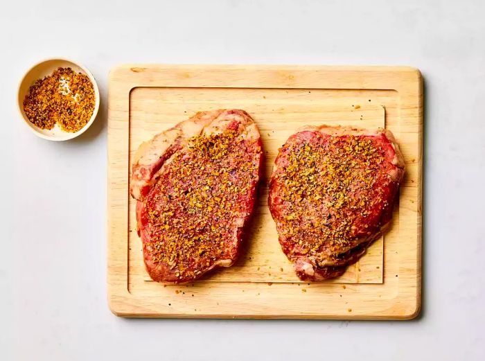 Two seasoned steaks resting on a cutting board with a bowl of seasoning beside them.