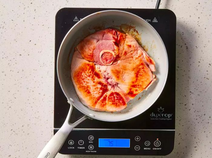 A golden-brown ham steak sizzling in a large skillet