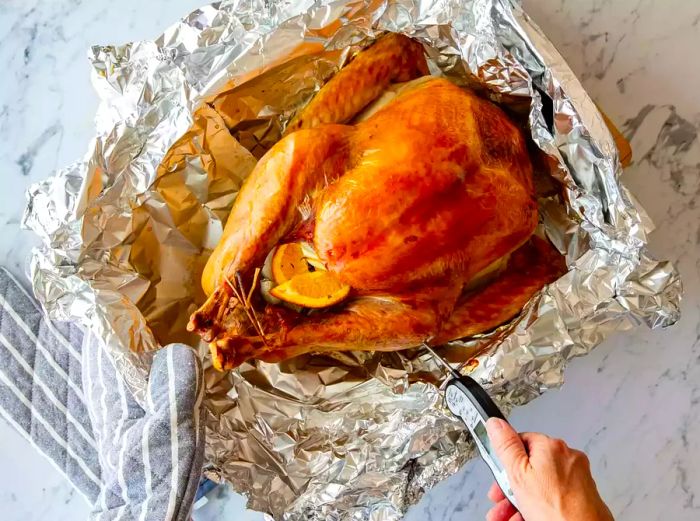 A person checking the internal temperature of a roasted turkey in a foil-lined roasting pan.