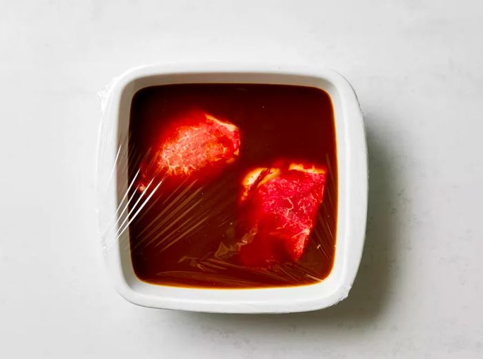 Steaks marinating in a white dish, covered with plastic wrap.