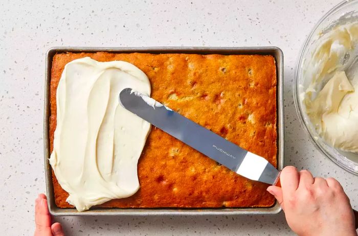 An offset spatula spreading a smooth layer of cream cheese frosting on a moist banana cake