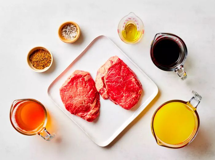 Two raw steaks with seasonings and ingredients arranged on a white countertop.