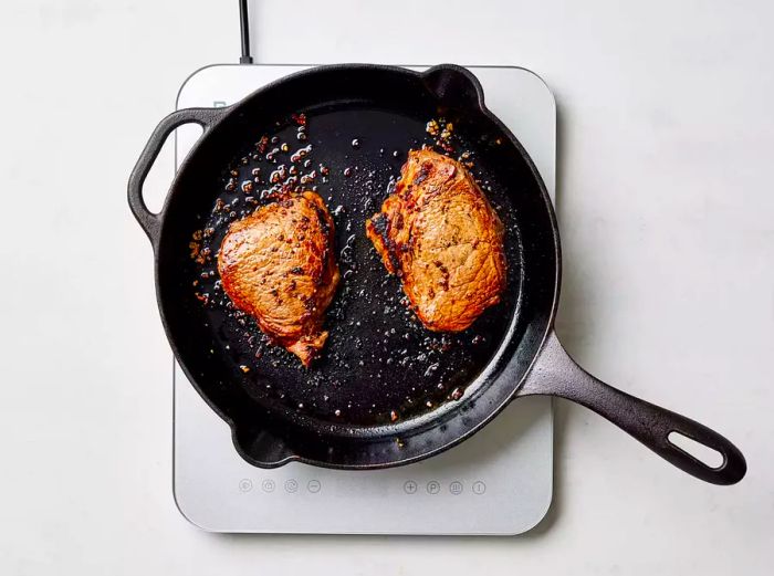 Steaks cooking in a cast-iron skillet, developing a brown crust on both sides.