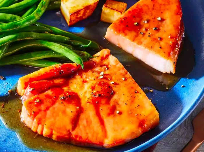 A plate featuring a sliced brown sugar ham steak, sprinkled with cracked black pepper, and accompanied by green beans and potatoes.