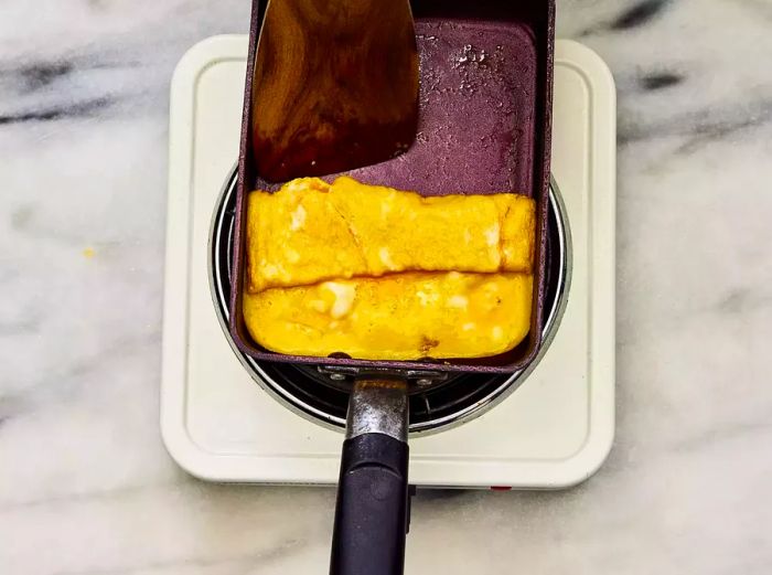 A spatula rolling a thin layer of cooked egg in a lightly greased omelet pan