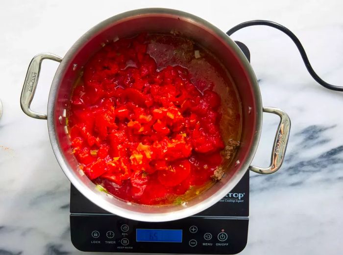 A large pot filled with seasoned ground beef, topped with stewed tomatoes, tomato sauce, diced tomatoes with green chiles, and water