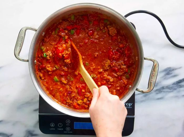 A wooden spoon mixing a large pot of beef and bean chili