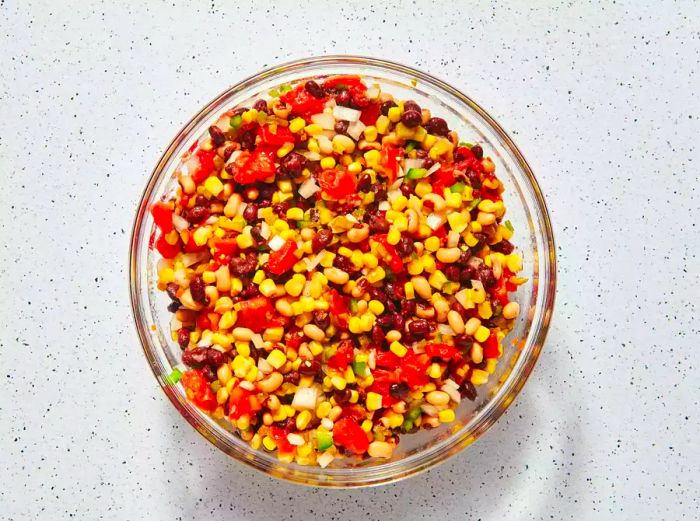 A glass bowl filled with black beans, black-eyed peas, diced tomatoes, corn, onions, bell peppers, and jalapeño peppers.