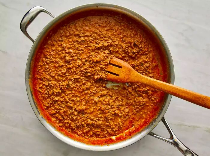 An overhead shot showing enchilada sauce and cheese being stirred into the ground beef and onion mixture in a large skillet.