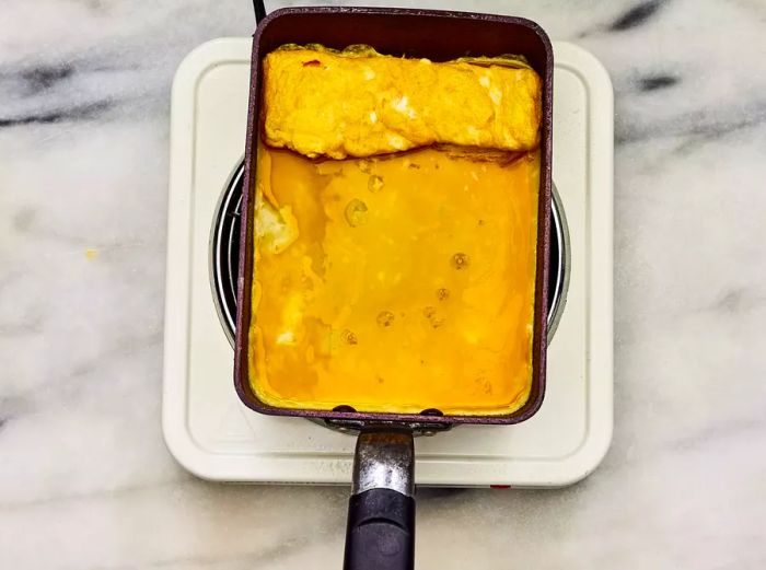 A rolled omelet at one end of the skillet, with more egg mixture poured into the pan