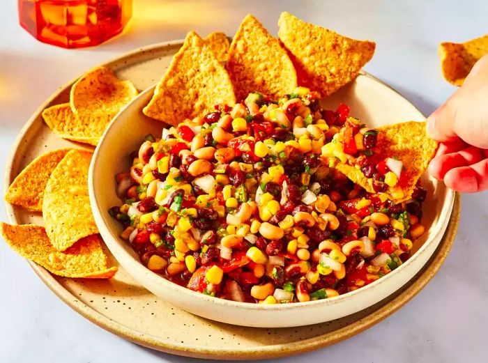 A bowl of cowboy caviar served alongside tortilla chips, with someone dipping a chip into the dip.