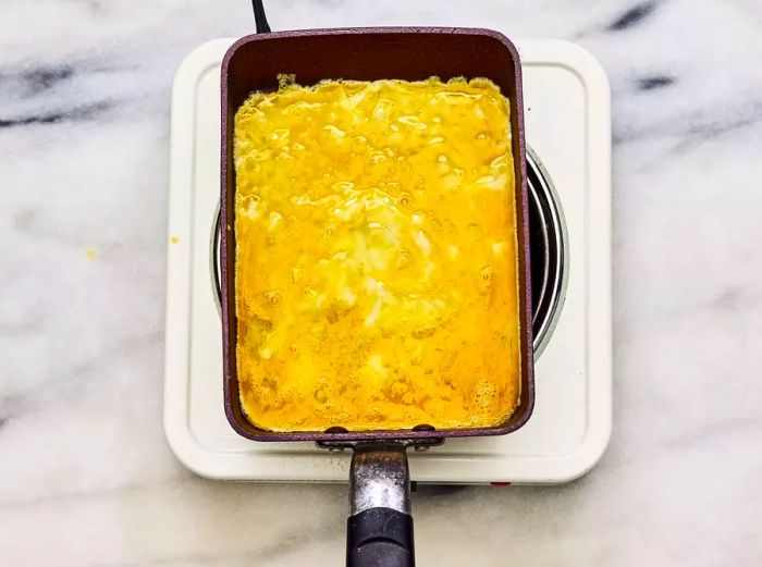 A greased nonstick omelet pan with a thin layer of egg mixture spread across the bottom