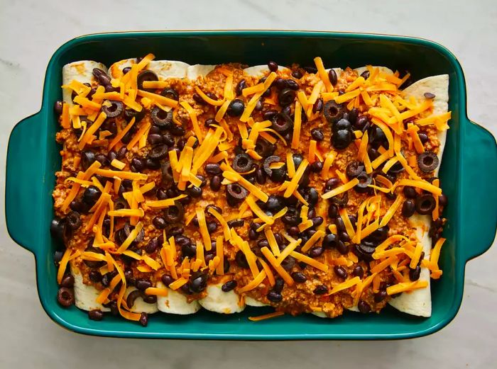 An overhead shot of an unbaked enchilada casserole, topped with sauce, sliced black olives, and a generous layer of shredded Cheddar cheese.