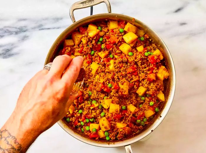 A hand sprinkling garam masala over a pan of Keema Aloo