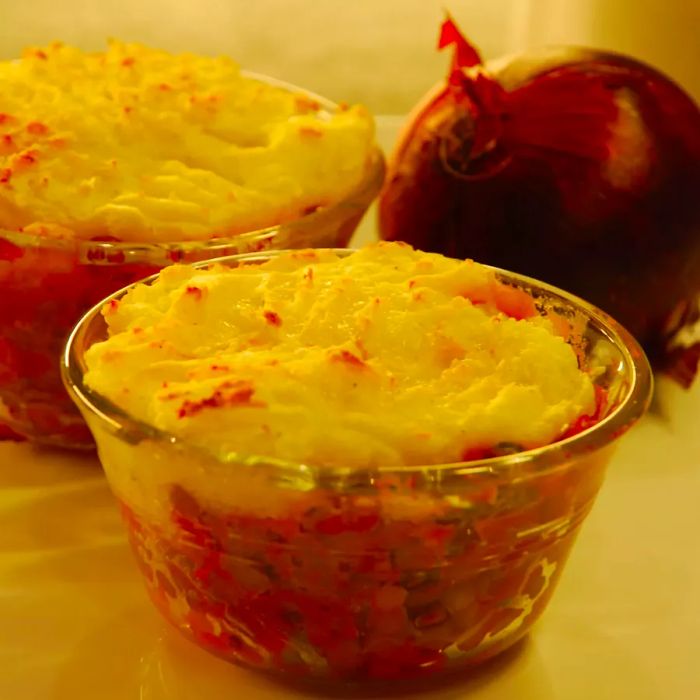 A close-up shot of individual servings of Vegan Shepherd's Pie in glass bowls, with a red onion in the background.