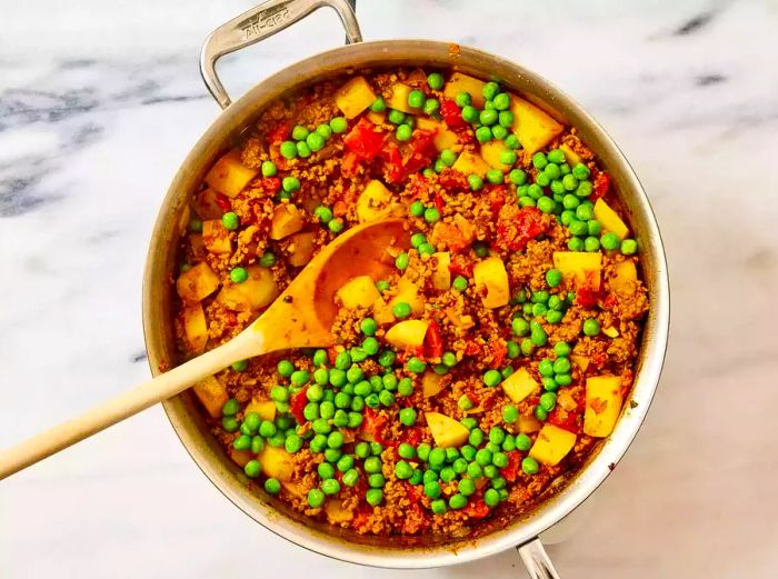 A skillet of ground beef mixed with tomatoes, potatoes, and peas, being stirred with a wooden spoon