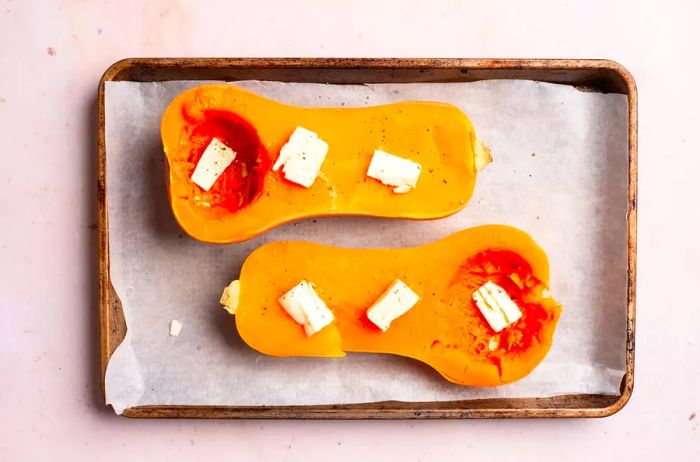 Butternut squash halved and placed on a parchment-lined baking sheet with pats of butter on top, ready for roasting.