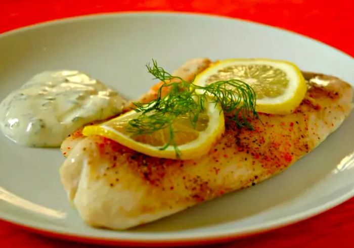 A baked tilapia fillet garnished with lemon slices and a sprig of fresh dill, served alongside tartar sauce.