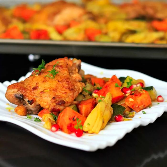 A square white plate featuring chicken and roasted vegetables, with a sheet pan of chicken in the background