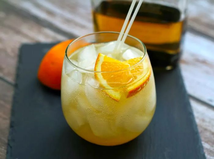 A rocks glass filled with an iced, fizzy drink made from golden tequila, garnished with orange slices, with an orange and tequila bottle in the background.