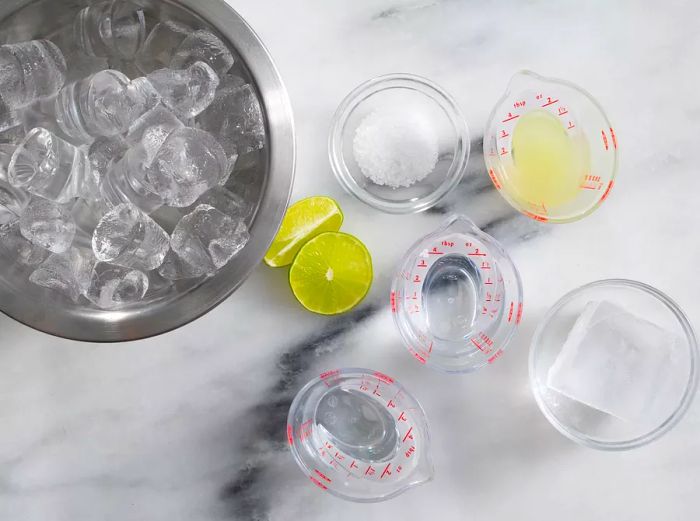 Aerial view of a bowl of ice with margarita ingredients in different measuring cups.